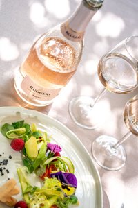 Ruggeri Argeo Rosé Flasche auf einem weißen Tisch, daneben zwei gefüllte Gläser. Im Vordergrund ein weißer Teller mit frischem Salat. Perfekte Kombination aus Roséwein und leichtem Gericht