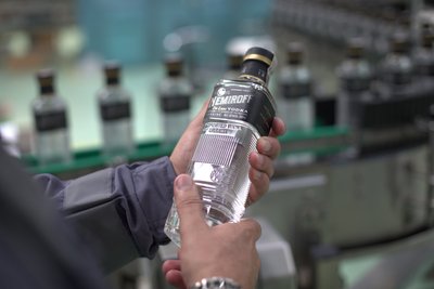 Eine Nemiroff DeLuxe Flasche wird in beiden Händen gehalten, während im Hintergrund ein Förderband mit weiteren Nemiroff Flaschen zu sehen ist