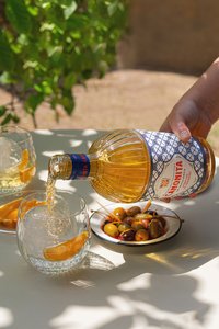 Canonita de Mallorca wird in ein Glas mit Eis und Orangenscheibe gefüllt. Auf dem Tisch stehen eine Schale mit grünen Oliven in Tomatensauce, eine Schale mit Orangenscheiben und ein weiteres Glas Canonita de Mallorca. Mediterraner Genuss und erfrischende Getränke perfekt präsentiert.