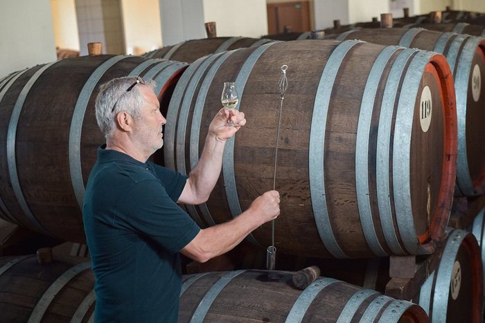Ein Mann überprüft die Qualität von Schnaps im Keller von Nordbrand Nordhausen, während im Hintergrund große Holzfässer stehen.