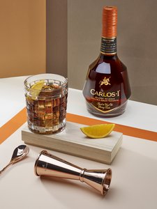 Auf einem Tisch steht eine Carlos I Flasche neben einem mit dem Brandy gefüllten Glas. Zusätzlich sieht man Cocktail Zubehör und eine aufgeschnittene Orange für den Drink. 