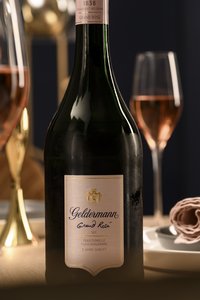 Nahaufnahme einer Geldermann Grand Rosé Sektflasche mit rosa Etikett. Im Hintergrund gefüllte Sektgläser.