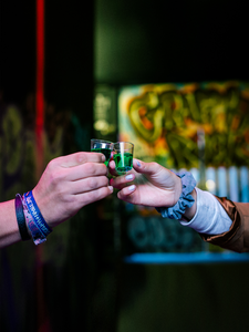 Das Bild zeigt zwei Hände, die grüne Shots mit Pfeffi halten und gerade anstoßen. Im Hintergrund erkennt man eine Bar, die eine lebendige Atmosphäre schafft.