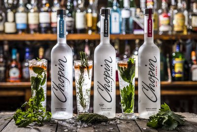Drei aufgereihte Chopin Vodka Flaschen mit den unterschiedlichen Sorten Roggen, Kartoffeln und Weizen. Dazu Gläser mit grünen Kräutern gefüllt. 