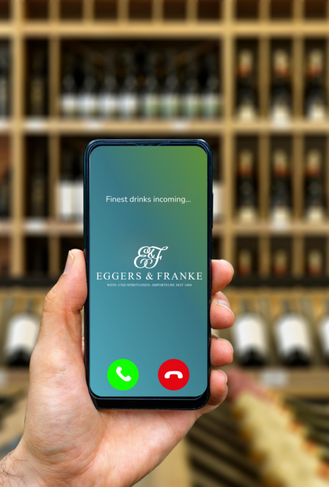 im Vordergrund hält jemand ein Handy in der Hand mit der Telefonnummer von Eggers und Franke Oesterreich, im Hintergrund sind Weinflaschen im Regal erkennbar